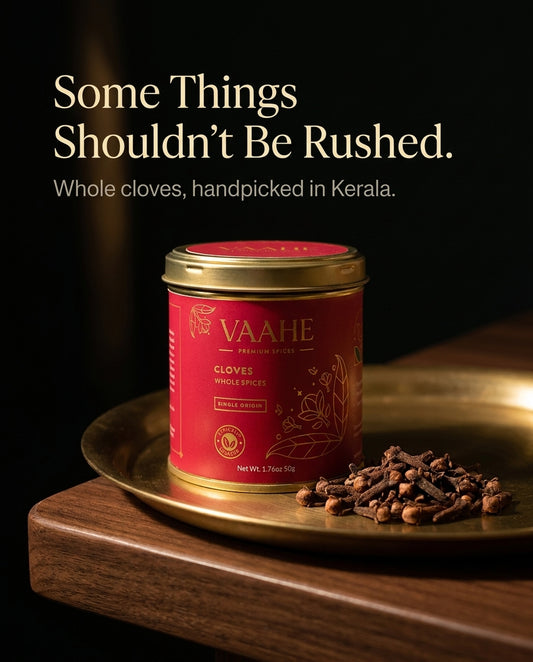 A red tin of VAAHE whole cloves sits on a brass plate with loose cloves beside it, set on a wooden surface against a dark background. Text at the top reads, 'Some Things Shouldn't Be Rushed.